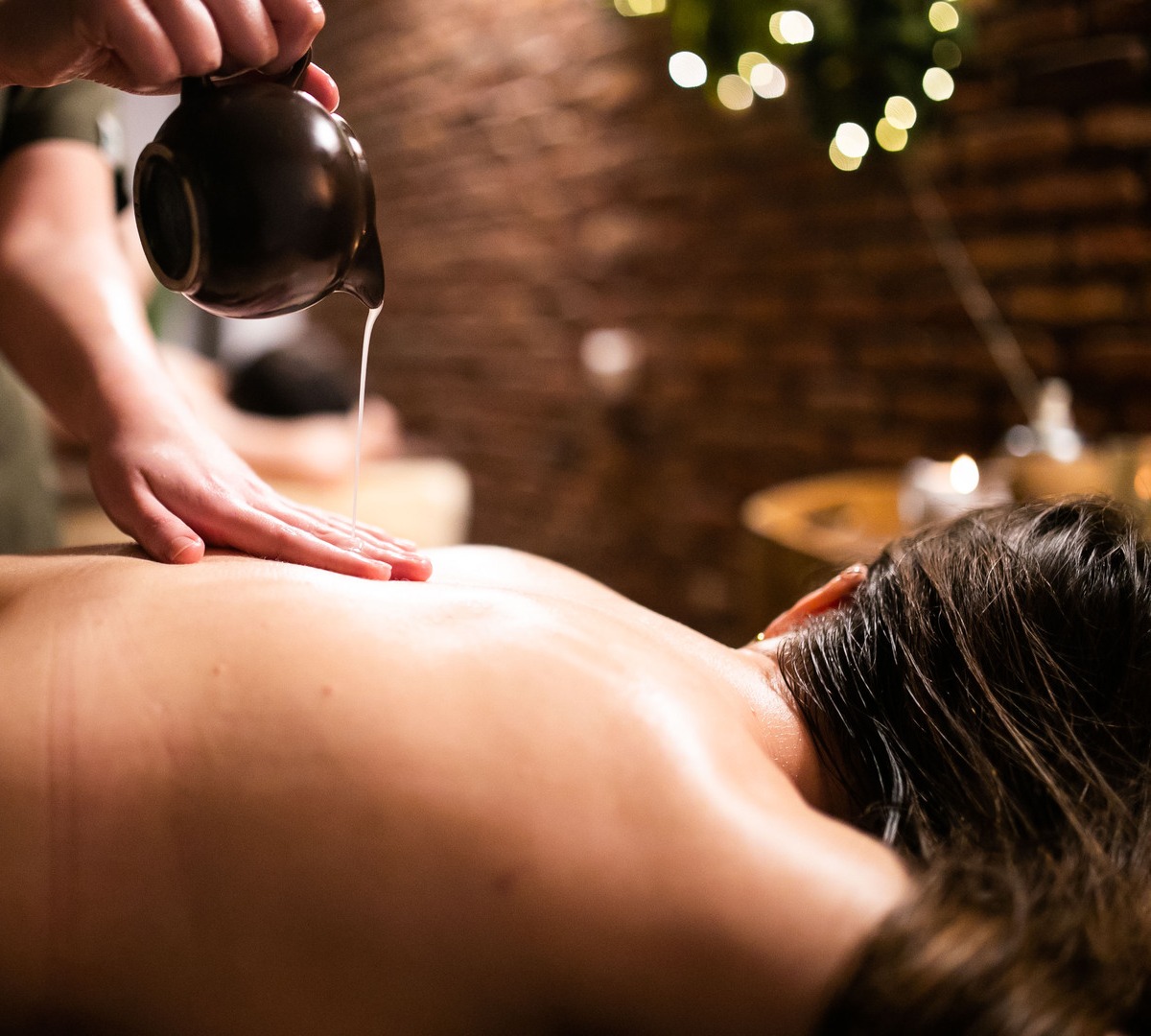 Spa therapist pouring warm oil and massaging a guest's bare back in a candlelit treatment room with exposed brick.