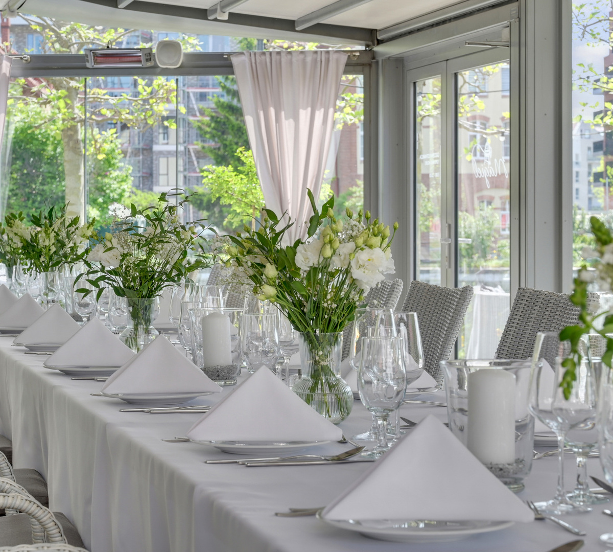 Langt bankettbord i glassveranda med hvit duk, brettede servietter, glass, hvite blomsteroppsatser og kurvstoler