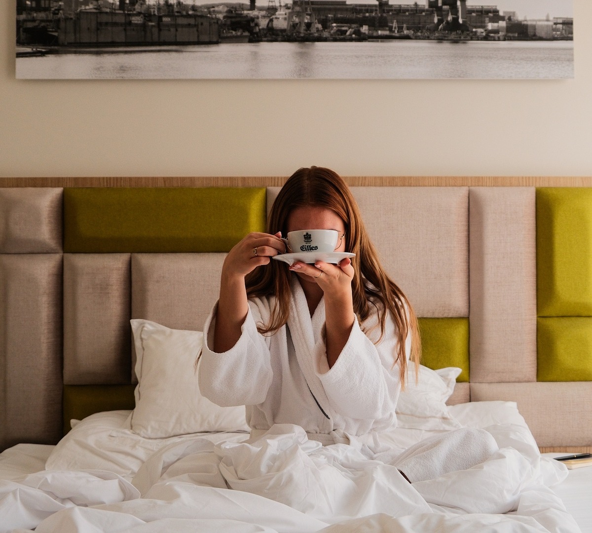 Gast im weißen Bademantel trinkt morgens Kaffee im Bett, weiches Kopfteil, frische Bettwäsche und gerahmtes Hafenfoto