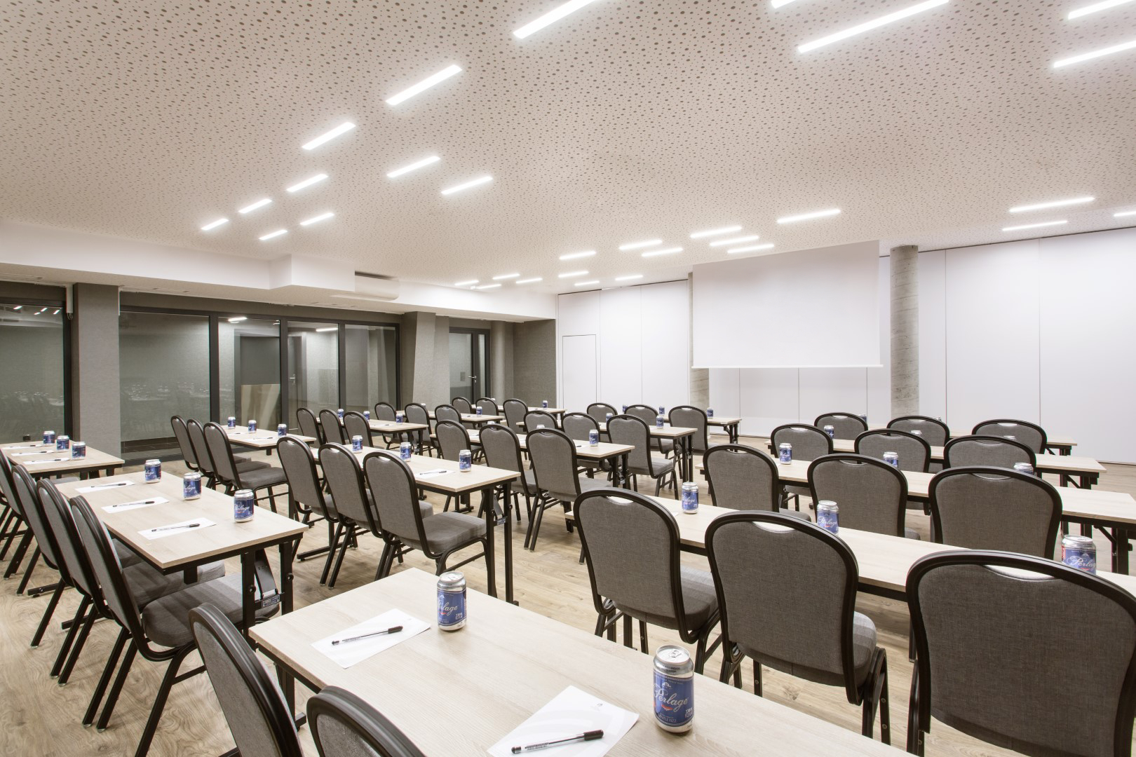 Moderne konferansesal på hotellet med klasseromsoppsett, rader med bord, penner, notatblokker, drikkebokser og projektorskjerm.