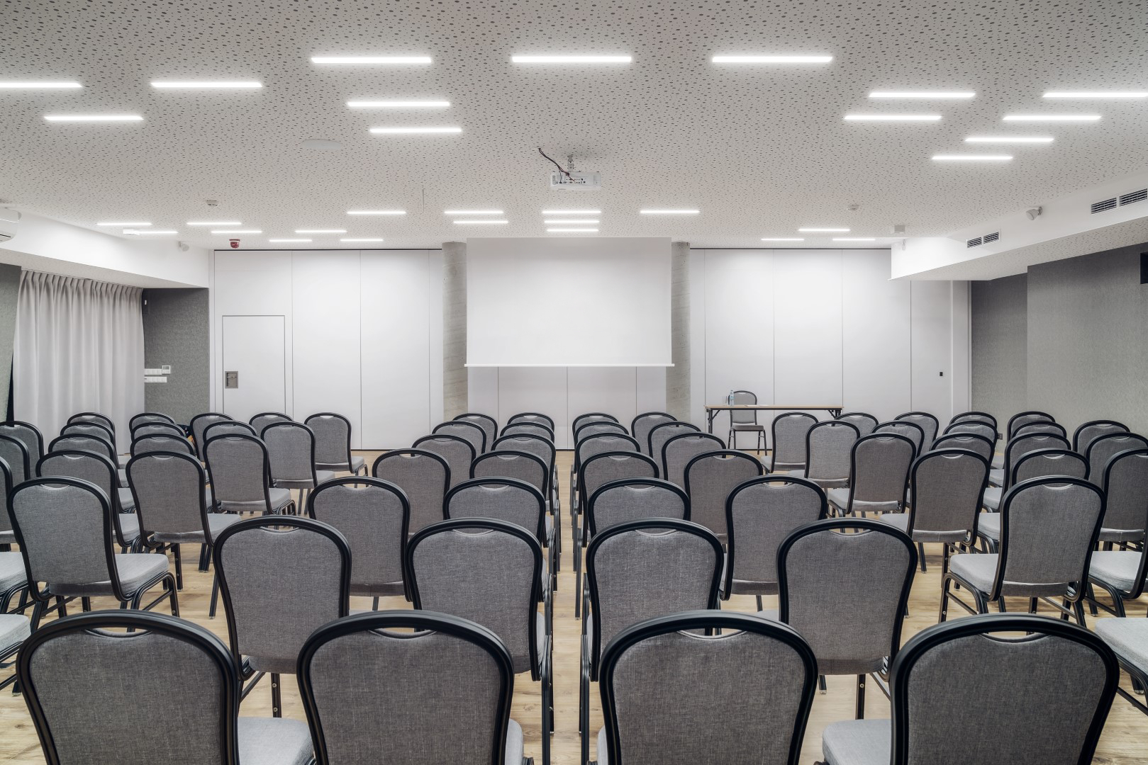 Moderne hotellkonferanserom med rader av grå stoler, projektorskjerm, foredragbord med vannflaske og dempet LED-belysning