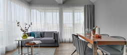 Jasny, nowoczesny apartamentowy salon z widocznym belkowaniem, zasłonami od podłogi do sufitu, szarą sofą, stolikami i stołem z szampanem
