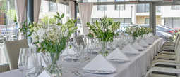Elegant lang bankettbord i glassveranda med hvite duker, blomsteroppsatser og rottingstoler