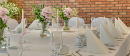 Bankettbord med hvit duk, brettede servietter, vinglass, rosa blomster i vaser og lys foran en murvegg