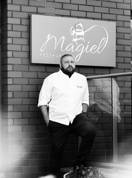 Head chef in white jacket leaning against brick wall outside Magiel restaurant by glass entrance and plants, relaxed.