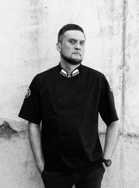 Male chef in black short-sleeve jacket standing against textured concrete wall, hands in pockets, serious expression