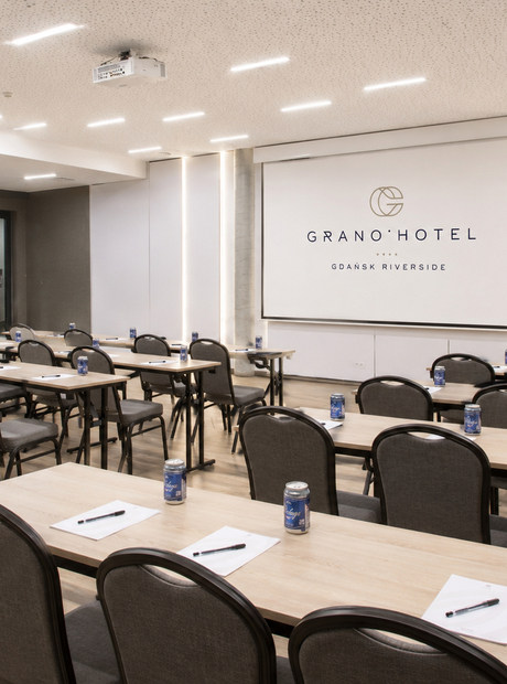 Moderner Hotelkonferenzraum im Klassenraumstil mit Projektor, Leinwand mit Grano Hotel‑Logo, Tische mit Schreibblöcken und Getränkedosen.