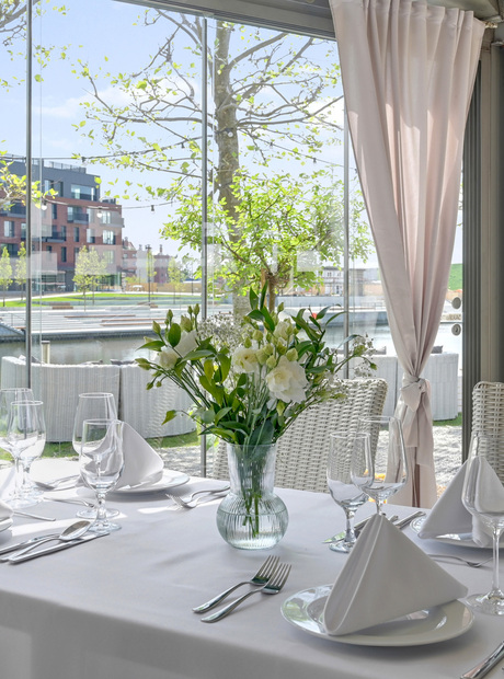 Lyst restaurantbord ved vannet med hvit duk, brettede servietter, glass og blomster, utsikt mot elva