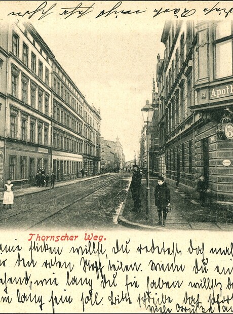 Zabytkowa uliczka z XIX‑wiecznymi fasadami, szyld apteki 'Apotheke', sklep z rumem i likierami, przechodnie, stara pocztówka.