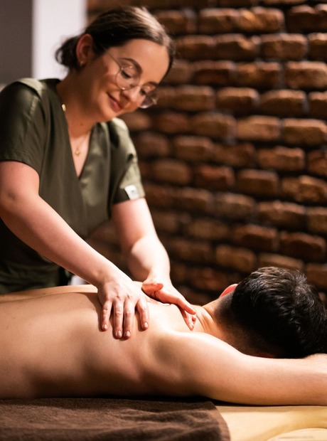 Terapeut ger avslappnande ryggmassage till en gäst i ett varmt spa med synlig tegelvägg och mjuka handdukar.