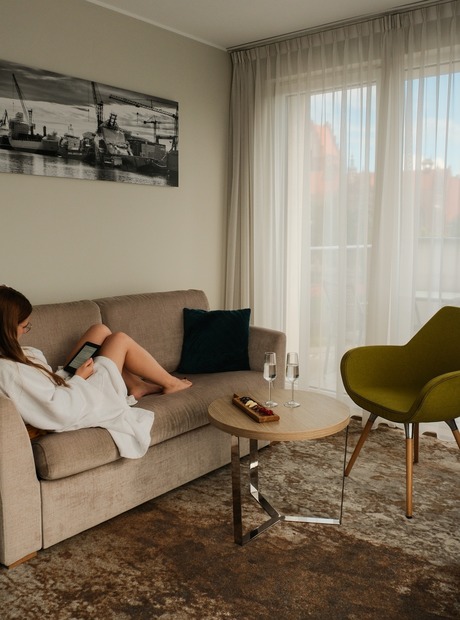 Wohnbereich der Suite mit beigefarbener Couch, Gast im Bademantel liest, helle Gardinen, Snackbrett und zwei Gläser