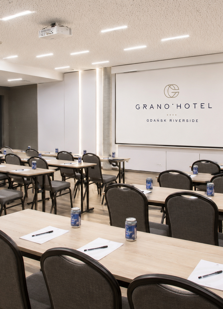 Nowoczesna sala konferencyjna w hotelu, ustawienie klasowe, projektor i ekran z logo Grano Hotel, stoły z notesami i napojami.