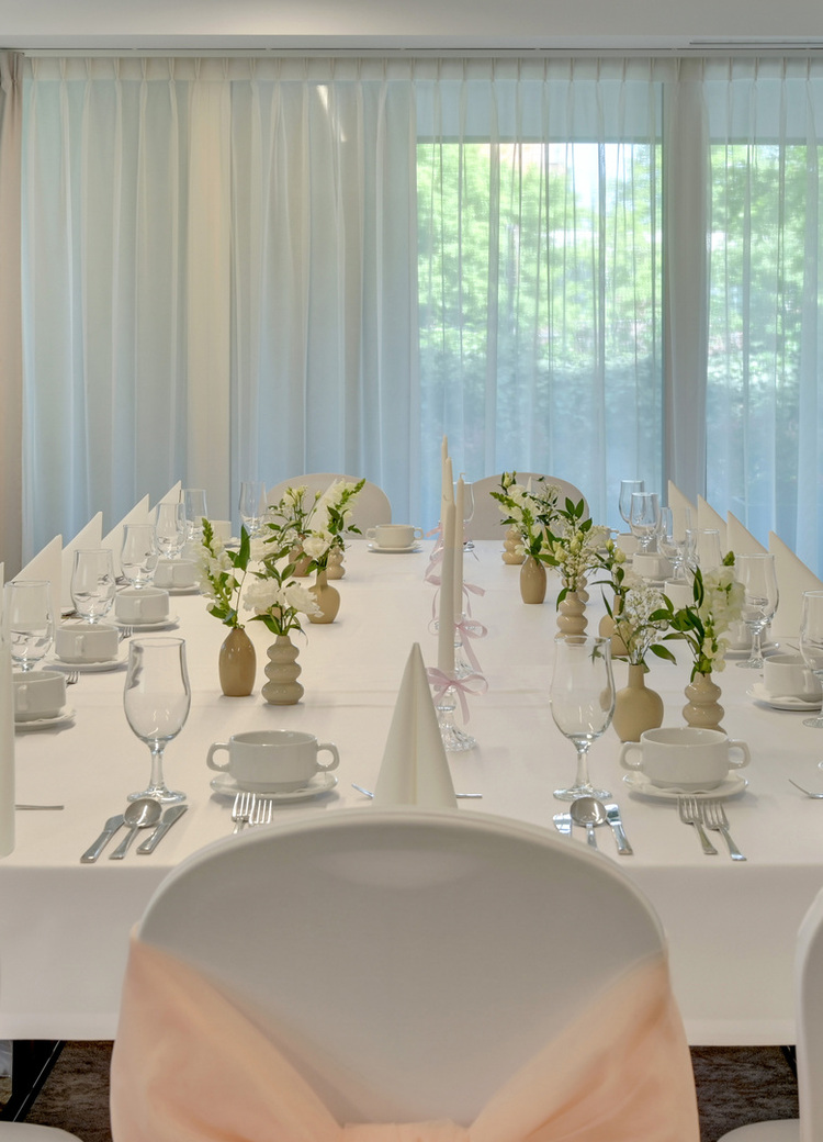Elegant bankettbord i lyst hotellrom, hvite duker, stoltrekk, brettede servietter, glass, blomsterdekorasjoner, naturlig lys