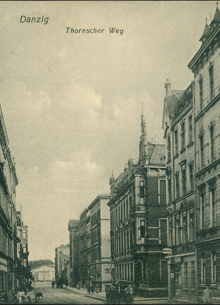Zabytkowa ulica z bogato zdobionymi elewacjami XIX w., torami tramwajowymi, narożną apteką z napisem 'Apotheke' i kilkoma przechodniami.