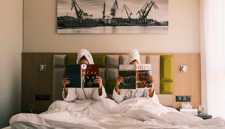Dwoje gości w szlafrokach z ręcznikowymi turbanami czyta magazyny na wygodnym hotelowym łóżku pod obrazem dźwigów