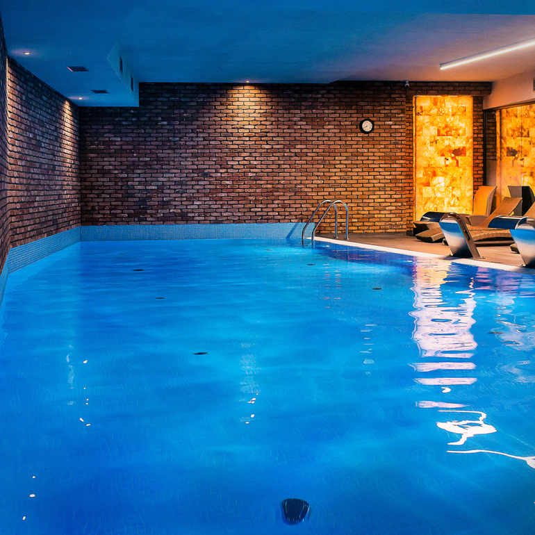 Uppvärmd inomhuspool på hotellet med blå LED-belysning, tegelvägg, metallstege och solstolar vid varm, bakbelyst saltstensvägg.