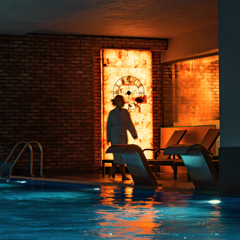 Inomhuspool med blått vatten, metallstege och solstolar, varm upplyst vägg med klockmotiv och silhuett av en gäst i morgonrock