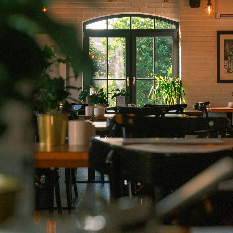 Przytulne wnętrze restauracji z łukowym oknem na bujny ogród, ciepłym oświetleniem i roślinami.