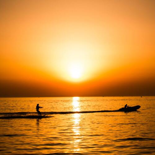 Gość uprawia narty wodne o zachodzie słońca na spokojnym morzu, ciągnięty przez motorówkę.