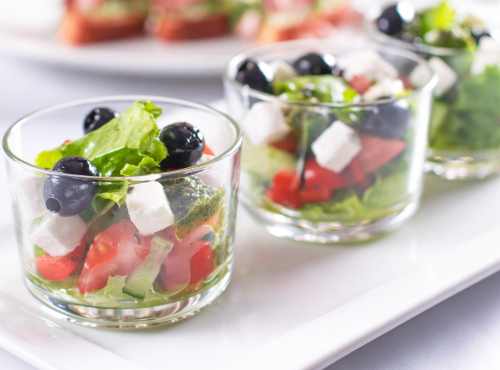 Elegancka przystawka w szklankach z sałatką grecką, oliwkami, serem feta i świeżymi warzywami na białym talerzu.