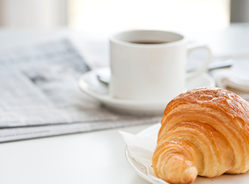 Świeżo upieczony rogalik na talerzu obok filiżanki kawy i gazet na jasnym stole w hotelu.