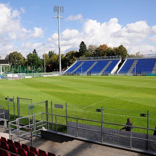Widok na piaszczystą murawę stadionu sportowego z trybunami i oświetleniem na tle nieba z chmurami.