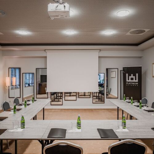 Nowoczesna sala konferencyjna z układem w podkowę, rzutnikiem, lustrami i wodą mineralną dla gości.