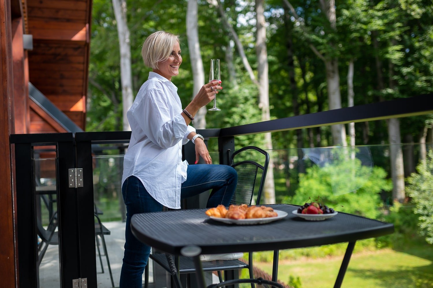 A smiling woman with a glass in her hand and her leg on a chair on a balcony with green background.