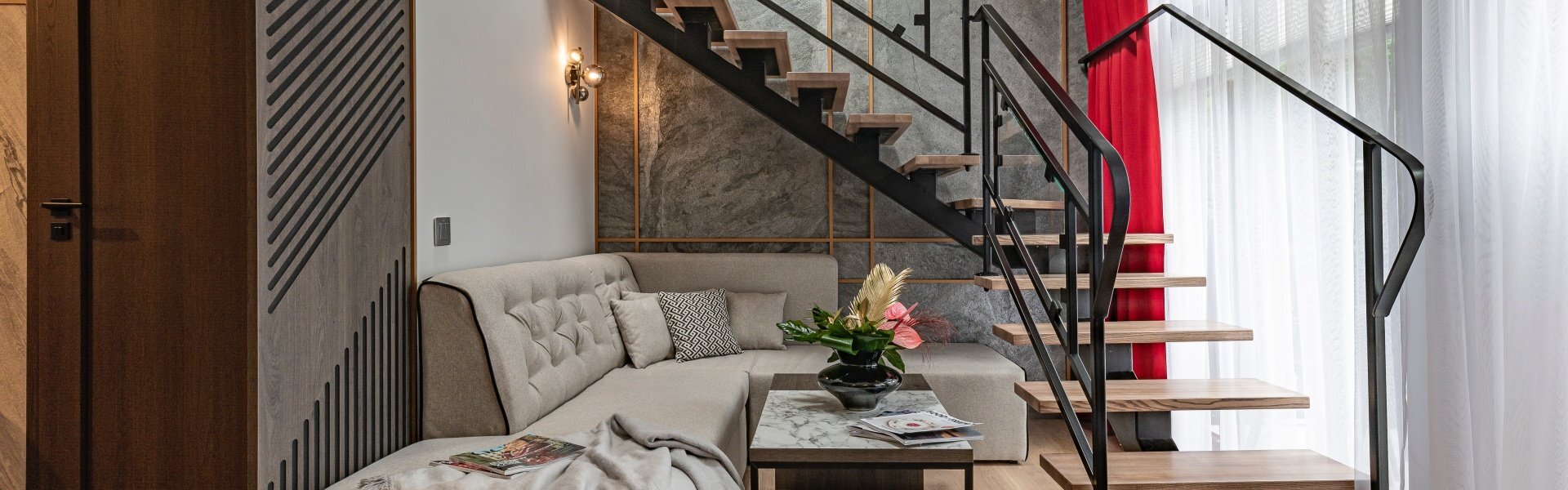 A high-ceilinged room with a light-colored corner sofa standing under wooden stairs with a dark rail