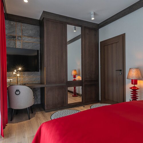 A room with a television on the wall, a desk and a chair next to a wardrobe with a mirror.