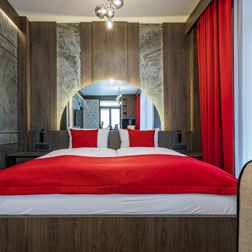 A double bed covered with a red bedspread and red cushions, and a window with a red curtain.