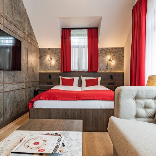 A hotel room with a double bed, red bedspread and a sofa next to a window with red curtains.