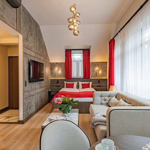 A living room with a sofa, a table and a bed in the background, by a window with red curtains.