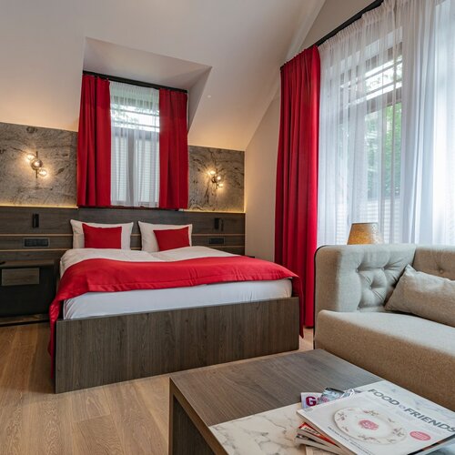 A hotel room with a double bed, red bedspread and a sofa next to a window with red curtains.