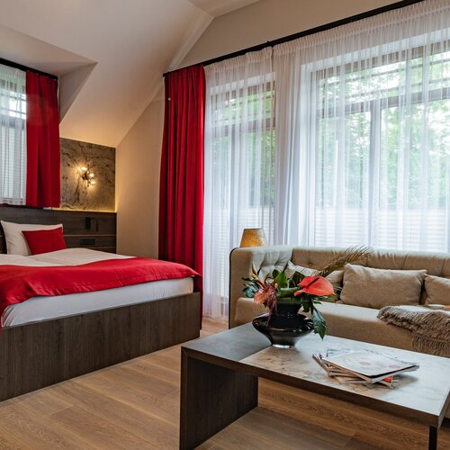 A room with a double bed, a sofa and a table with flowers by a large window.