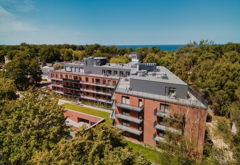 Nowoczesny hotel z czerwonej cegły z balkonami, otoczony zielenią, z widokiem na morze i las w tle.