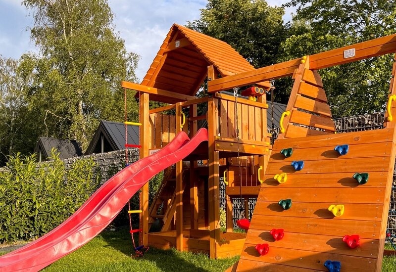 Drewniany plac zabaw z czerwonym zjeżdżalnią i ścianką wspinaczkową w ogrodzie hotelu, otoczony zielenią.