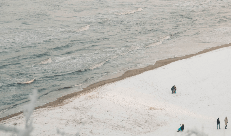 Winterlicher Strand mit Schneebedeckung, einige Personen spazieren und spielen am ruhigen Meeresstrand.