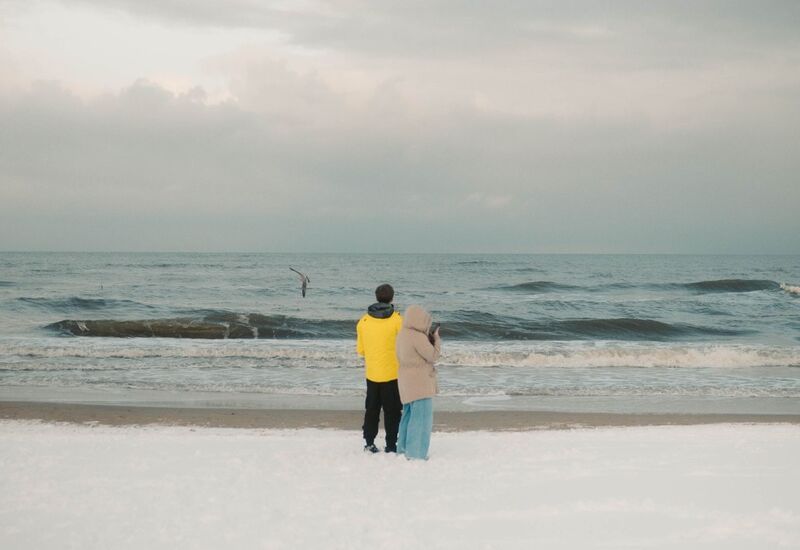 Para w ciepłych ubraniach stoi na śnieżnej plaży i podziwia zimne fale morza pod pochmurnym niebem.