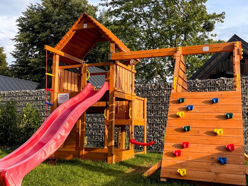Drewniany plac zabaw z czerwonym zjeżdżalnią i ścianką wspinaczkową w hotelowym ogrodzie na trawniku