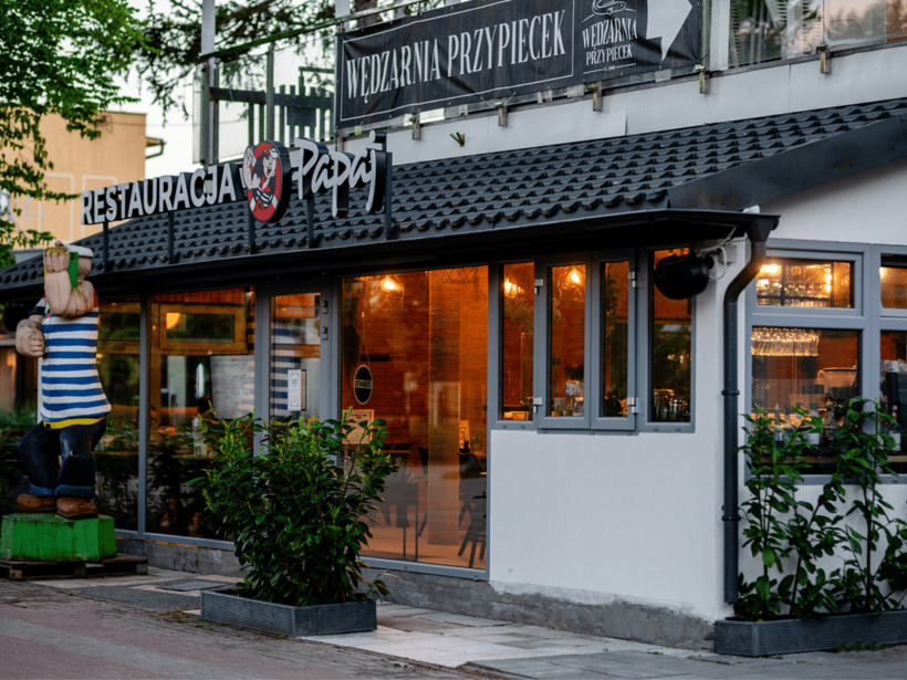 Wejście do restauracji o przytulnym wnętrzu, szklane okna i drewniana rzeźba marynarza na zewnątrz