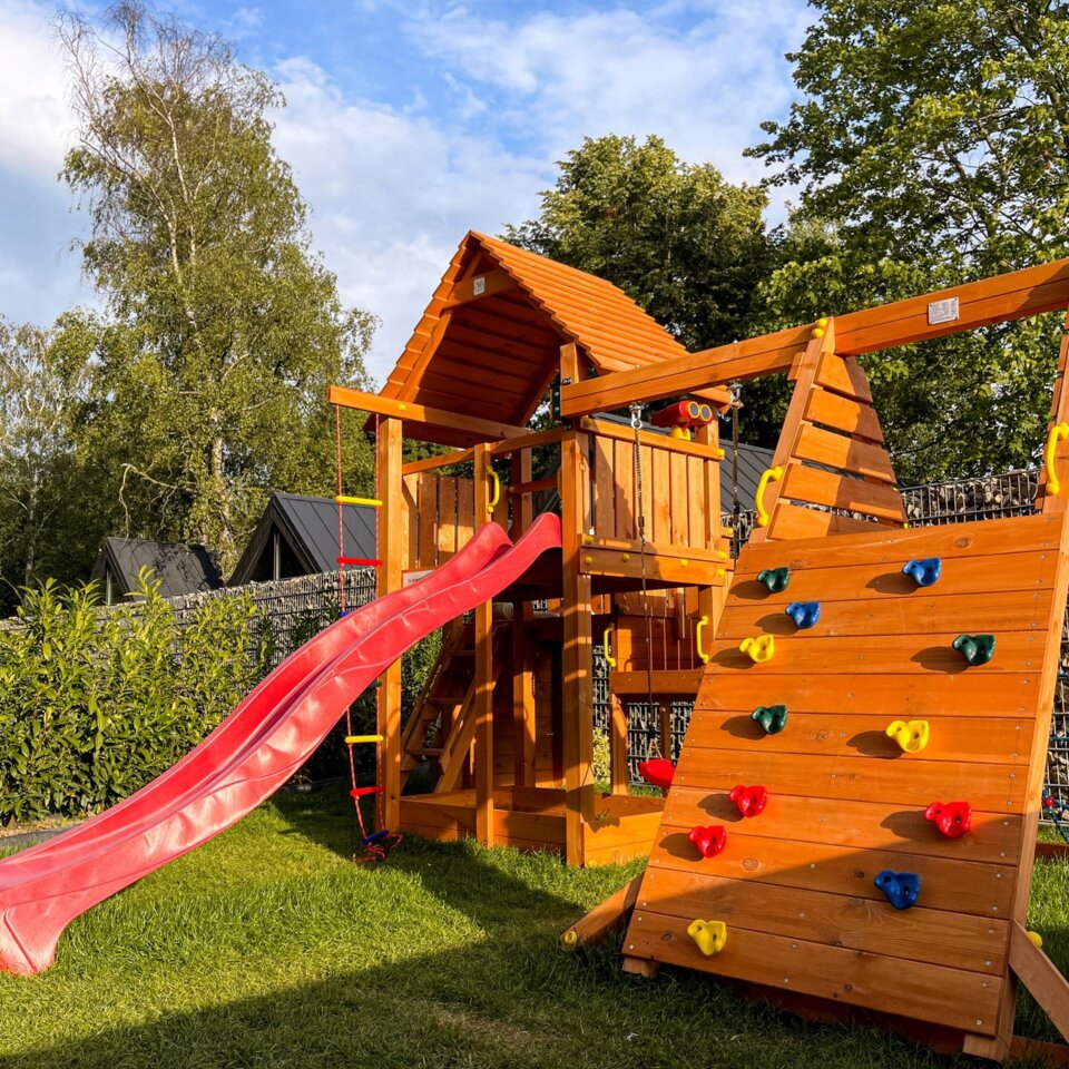 Holzspielplatz mit roter Rutsche und Kletterwand im Hotelgarten, umgeben von üppigem Grün und Bäumen.