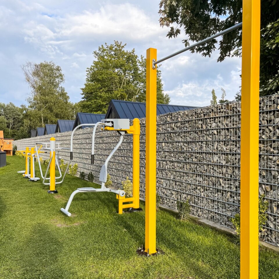 Gelbe Außenfitnessgeräte auf Rasenfläche neben Steinmauer, umgeben von Bäumen und Grünflächen.