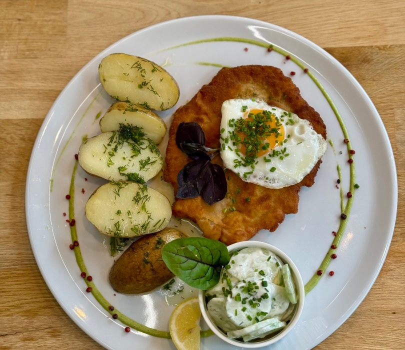 Chrupiący kotlet z sadzonym jajkiem, ziemniakami z koperkiem i sosem ogórkowym na eleganckim białym talerzu.