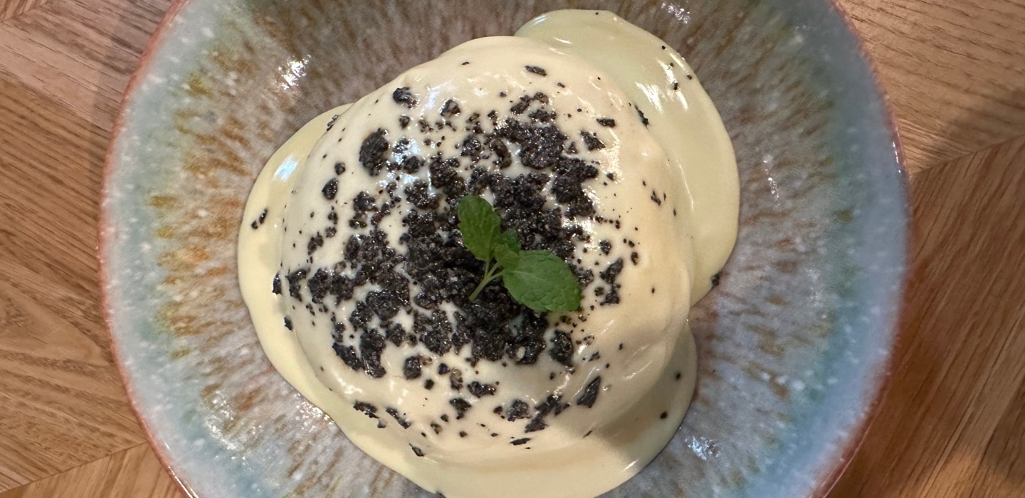 Delikatny deser z kremową polewą, posypany ciemnymi okruszkami i ozdobiony listkiem mięty na ceramicznym talerzu