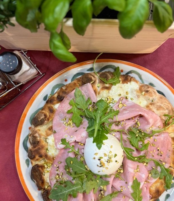 Pizza z grillowanym ciastem, szynką, rukolą i kulką sera burrata na ceramicznym talerzu przy świeżej bazylii.