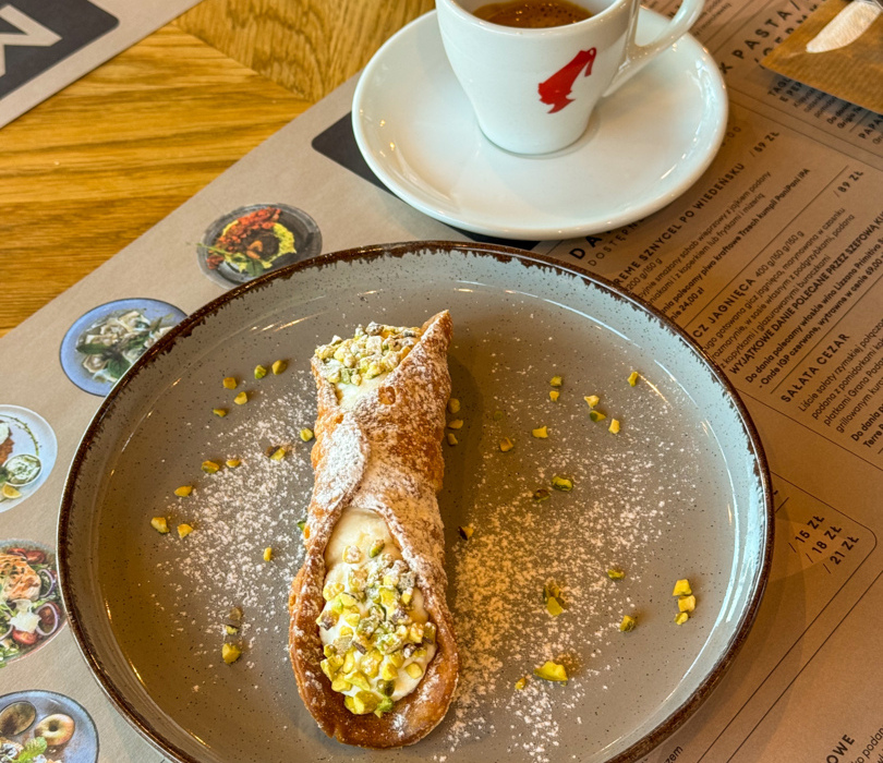 Deser cannoli z kremem ricotta i pistacjami podany na ceramicznym talerzu, obok filiżanka kawy espresso Julius Meinl