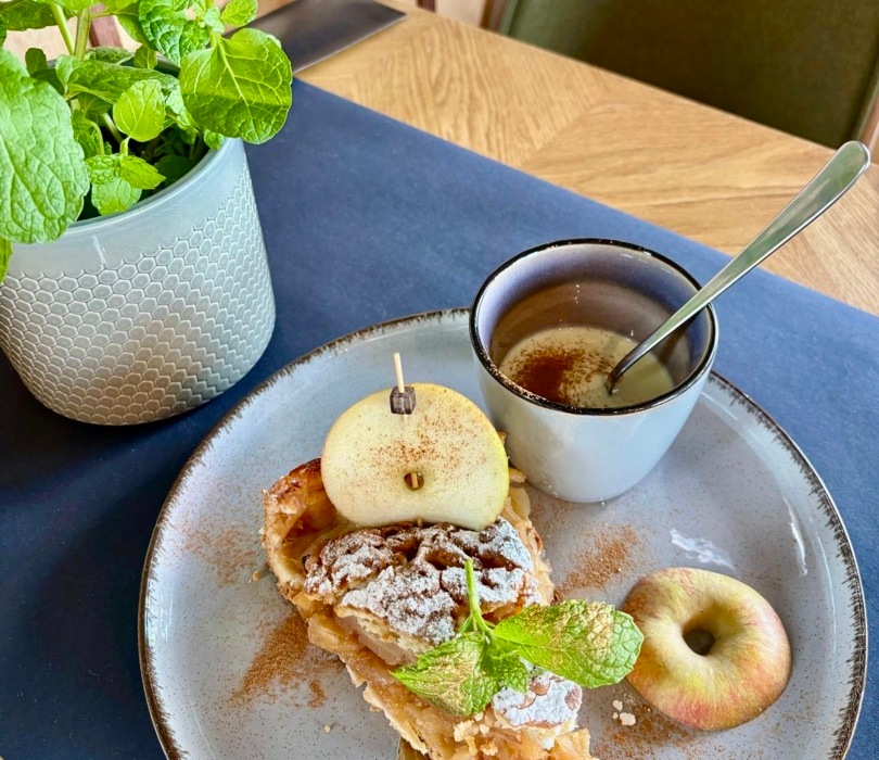 Plasterek szarlotki z cukrem pudrem i miętą na talerzu z jabłkiem i filiżanką herbaty na drewnianym stole