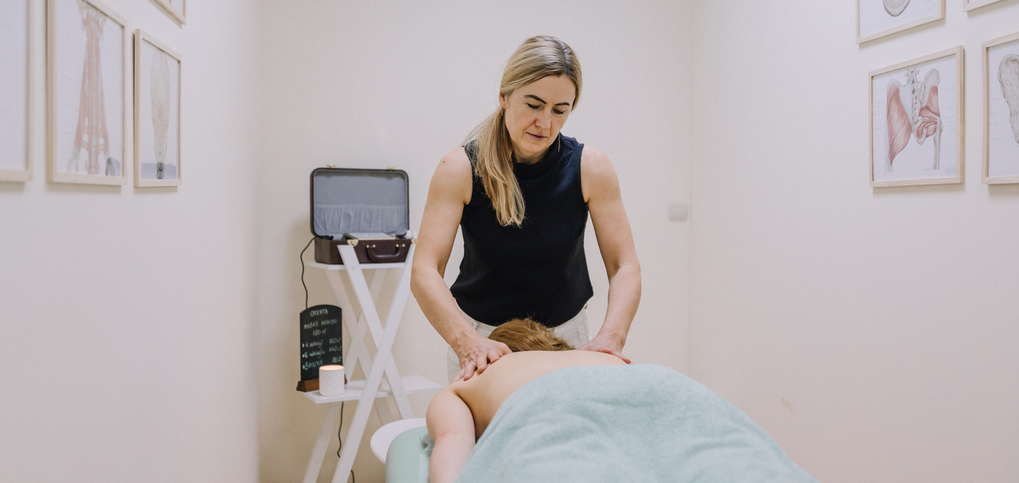 Therapeutin gibt Rückenmassage in ruhigem Spa-Behandlungsraum, weiches Licht, Gast unter Handtuch und anatomische Wandbilder