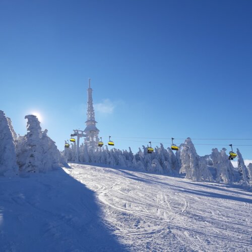 Zimowy stok narciarski pokryty śniegiem, wyciąg krzesełkowy z żółtymi siedzeniami i wieża radiowa na tle jasnego, błękitnego nieba.